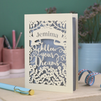 Decorative papercut graduation card with 'Jemima Follow Your Dreams' text on a desk with stationery items.
