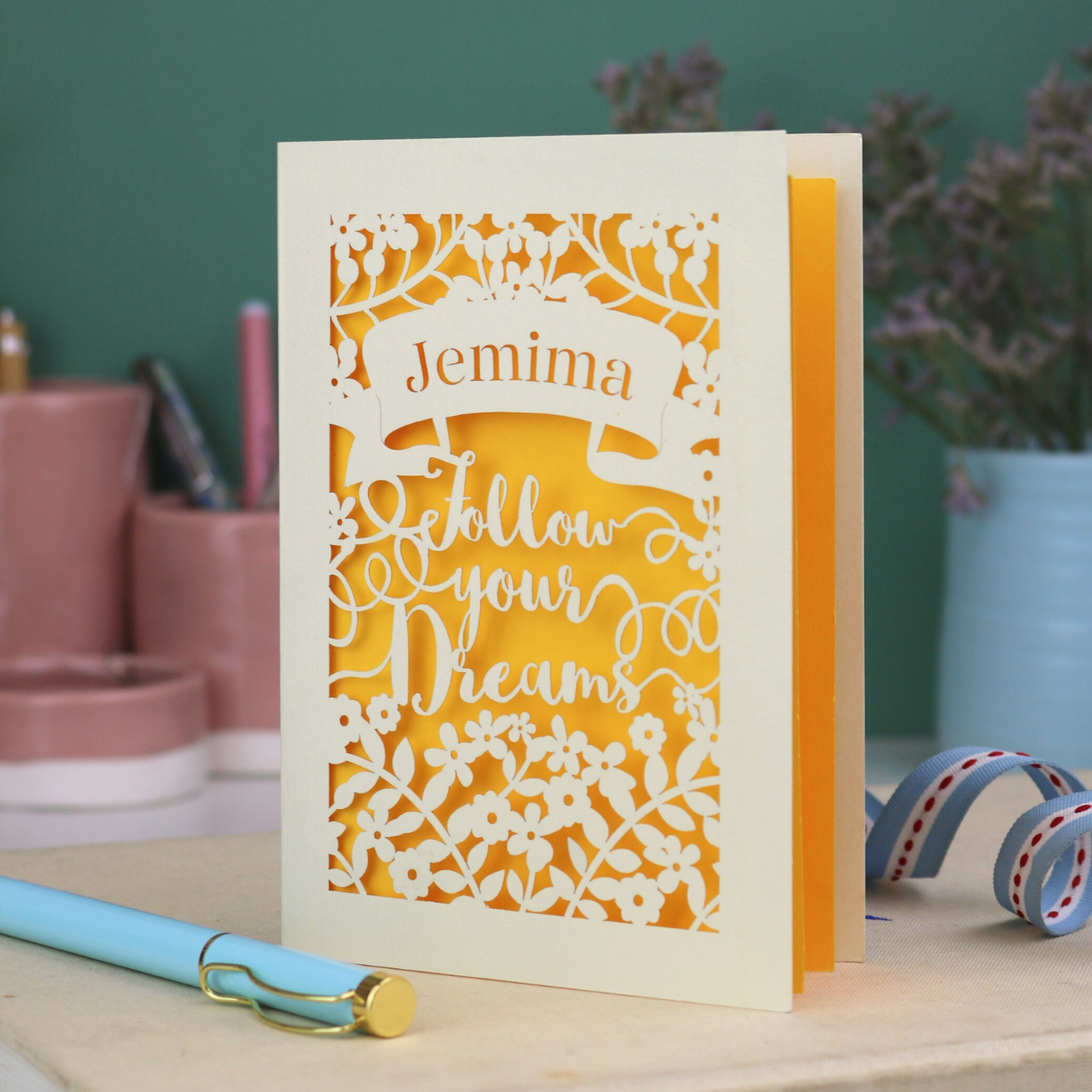 Decorative papercut graduation card with 'Jemima Follow Your Dreams' text on a desk with stationery items.