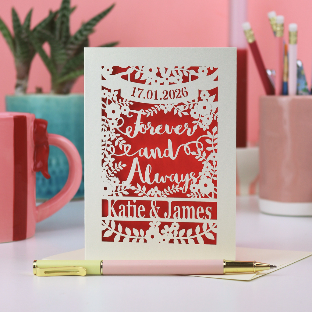 A laser cut engagement card with a date and the words "Forever and Always" with names and floral design. Card is on a white desk with stationery items and a pink background