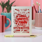 A laser cut engagement card with a date and the words "Forever and Always" with names and floral design. Card is on a white desk with stationery items and a pink background
