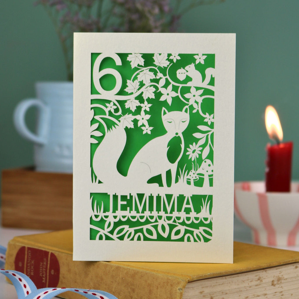 A laser cut birthday card with woodland scene and fox cut out design, personalised with an age in the top left, and a name at the bottom. 