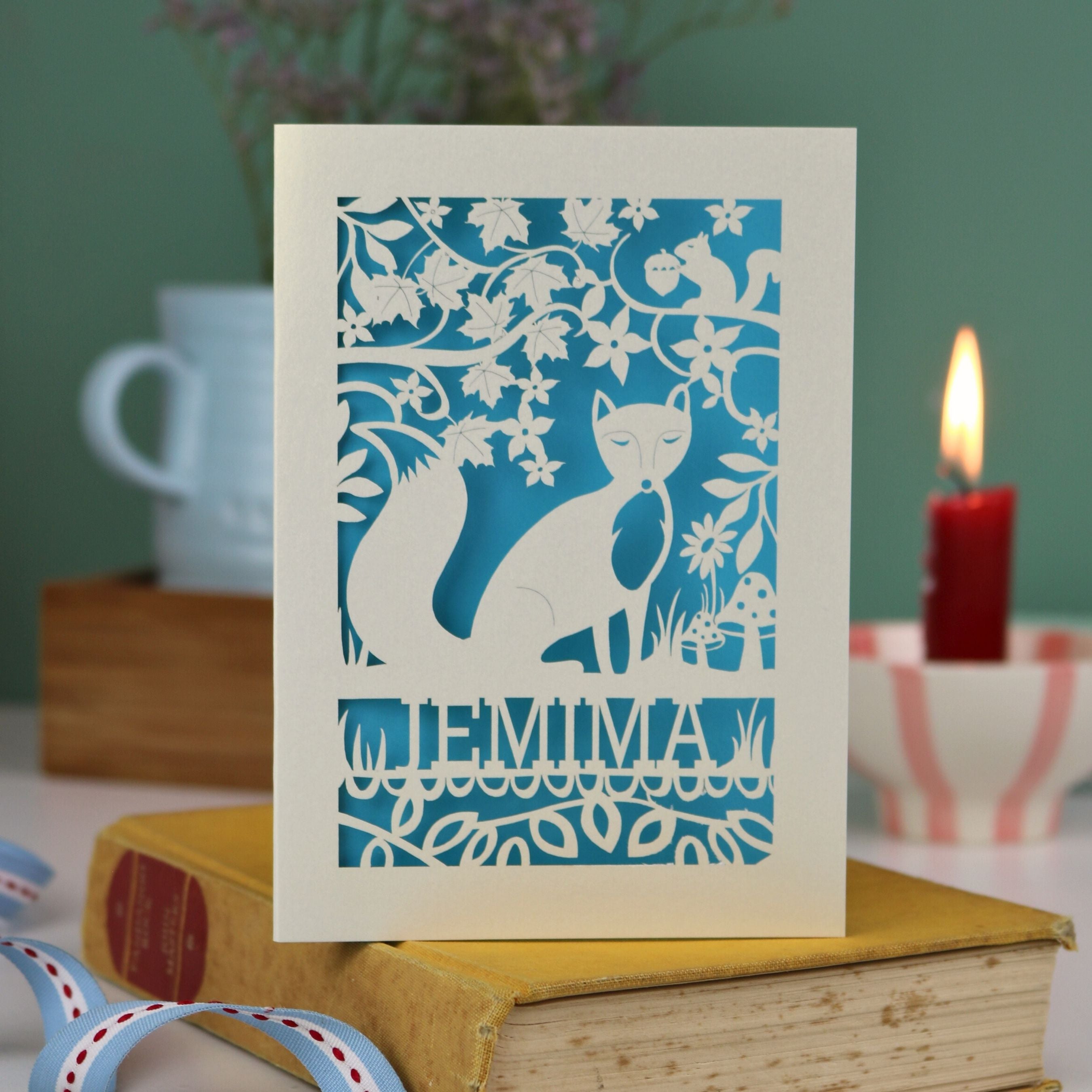 A laser cut fox birthday card with cut out fox illustration and woodland details, personalised with a name underneath the design. 