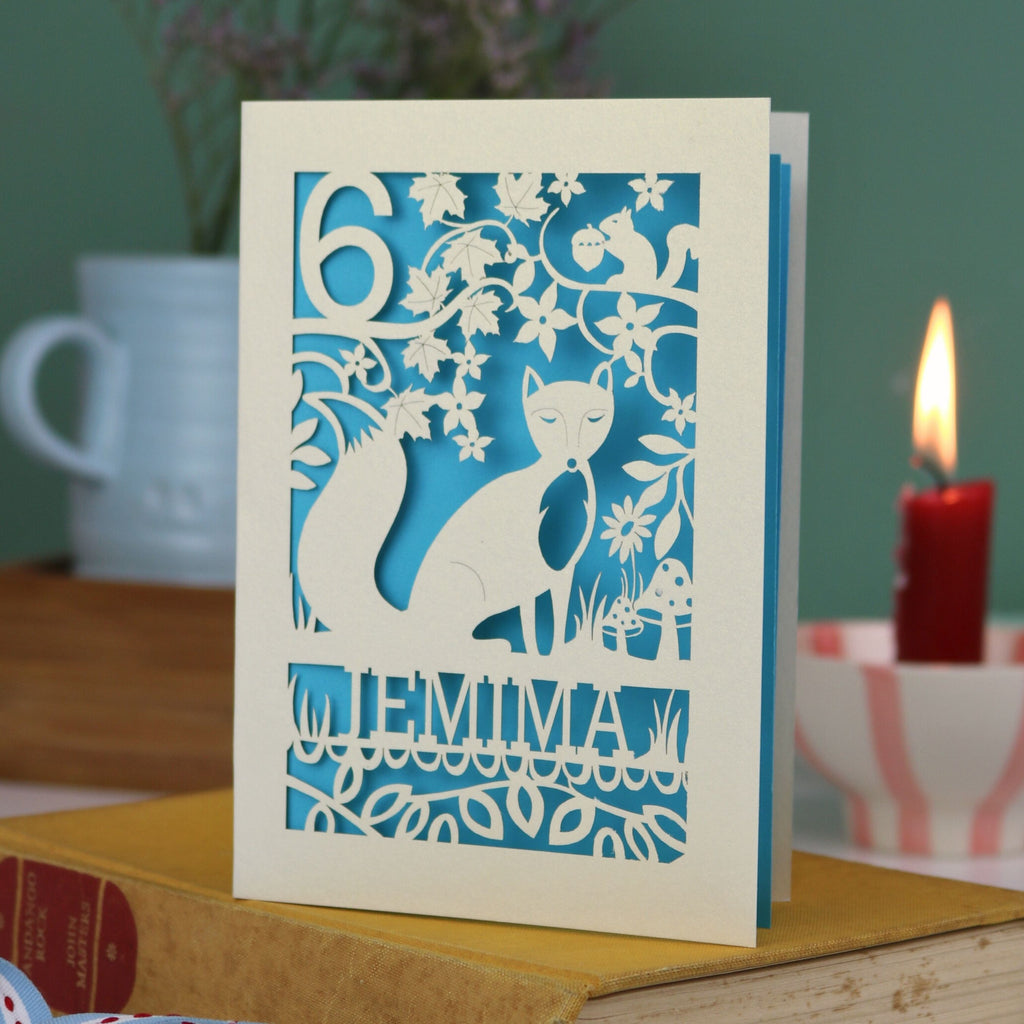 A laser cut birthday card with woodland scene and fox cut out design, personalised with an age in the top left, and a name at the bottom. 