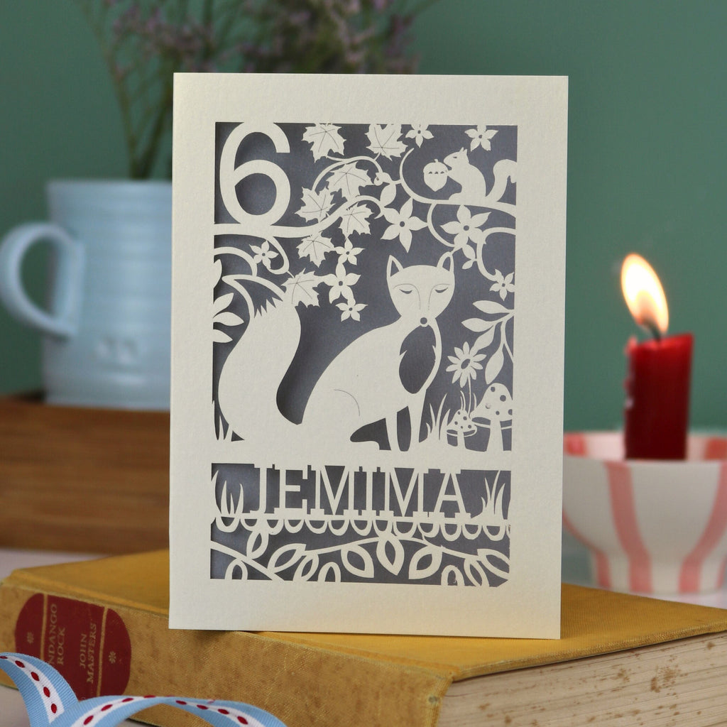 A laser cut birthday card with woodland scene and fox cut out design, personalised with an age in the top left, and a name at the bottom. 