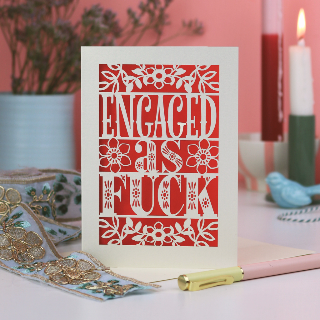 A laser cut funny engagement card that says "Engaged as fuck", on a white desk with stationery items and a pink background