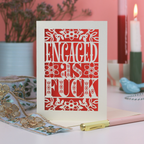 A laser cut funny engagement card that says "Engaged as fuck", on a white desk with stationery items and a pink background