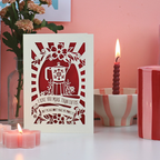 A funny laser cut Valentine's card on desk with candles and flowers, and a pink background