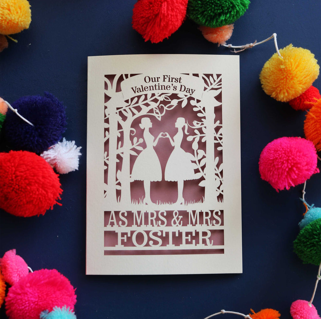 A personalised Our first Valentine's day card with a laser cut design of two women making a heart shape, with a blank space for a personal message, and brightly coloured pom-pom decorations around it.