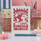 A laser cut girlfriend Valentine's card with globe design on a desk with stationery items and candles, and a pink background.