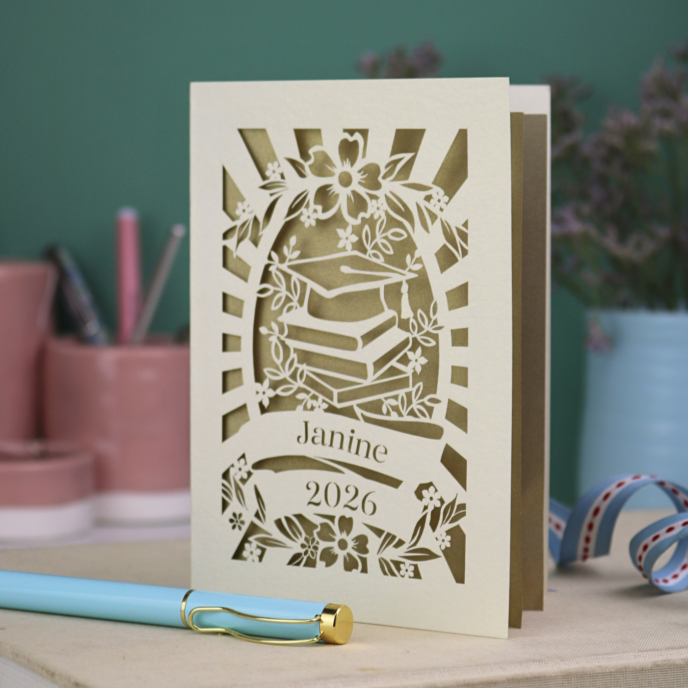 Personalised graduation card with cap and book design, and personalised with a name and the year in stencil font in a banner