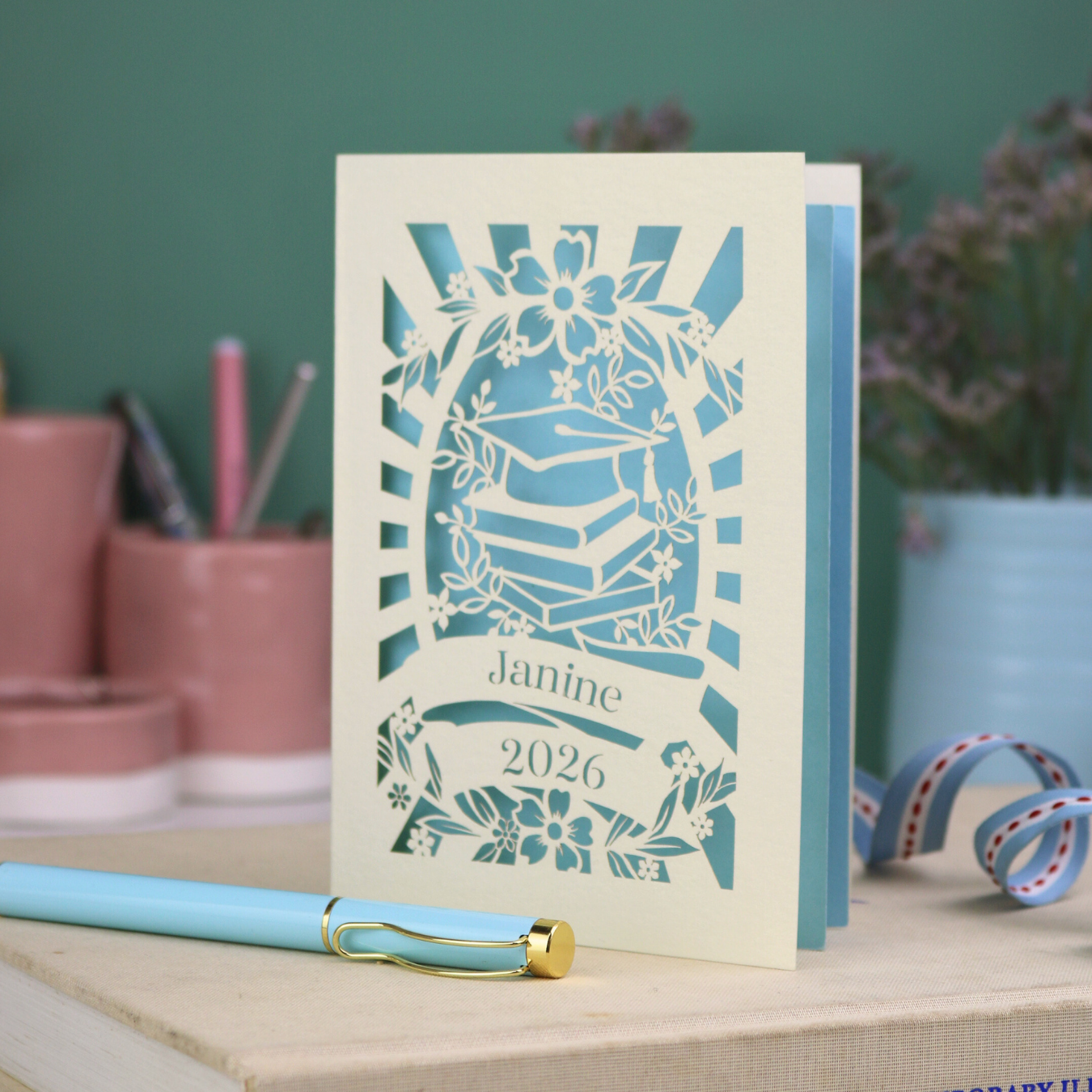 Personalised graduation card with cap and book design, and personalised with a name and the year in stencil font in a banner