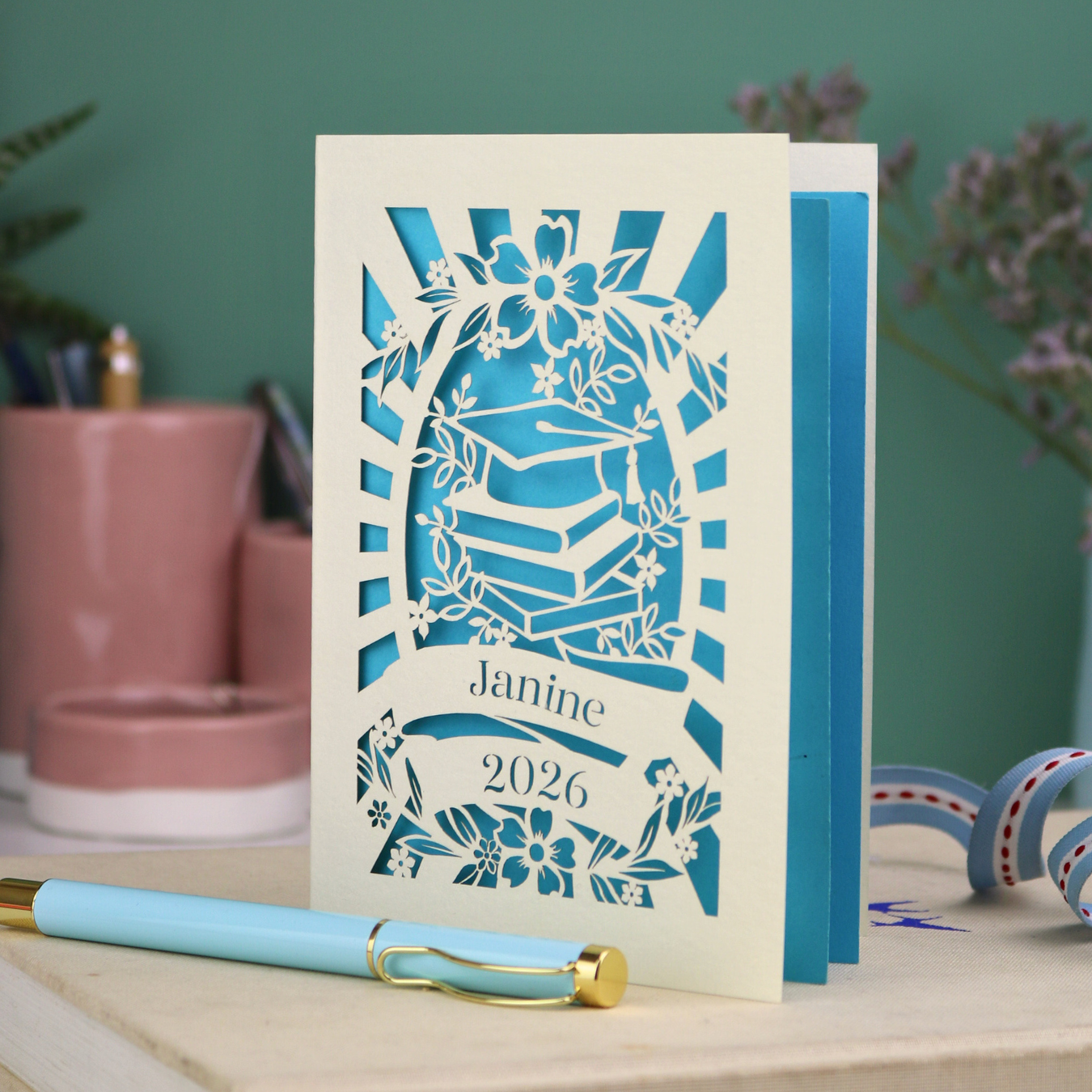 Personalised graduation card with cap and book design, and personalised with a name and the year in stencil font in a banner