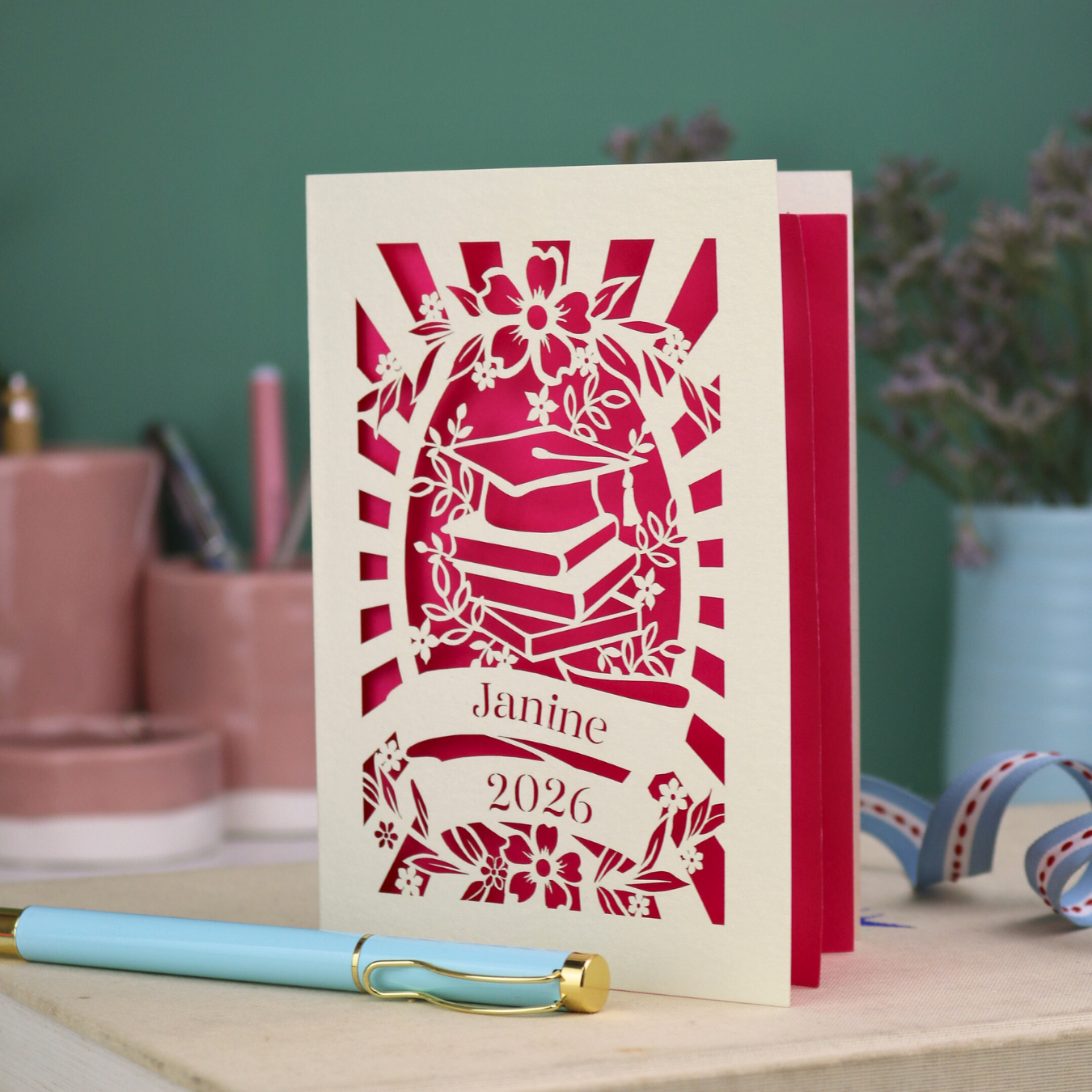 Personalised graduation card with cap and book design, and personalised with a name and the year in stencil font in a banner