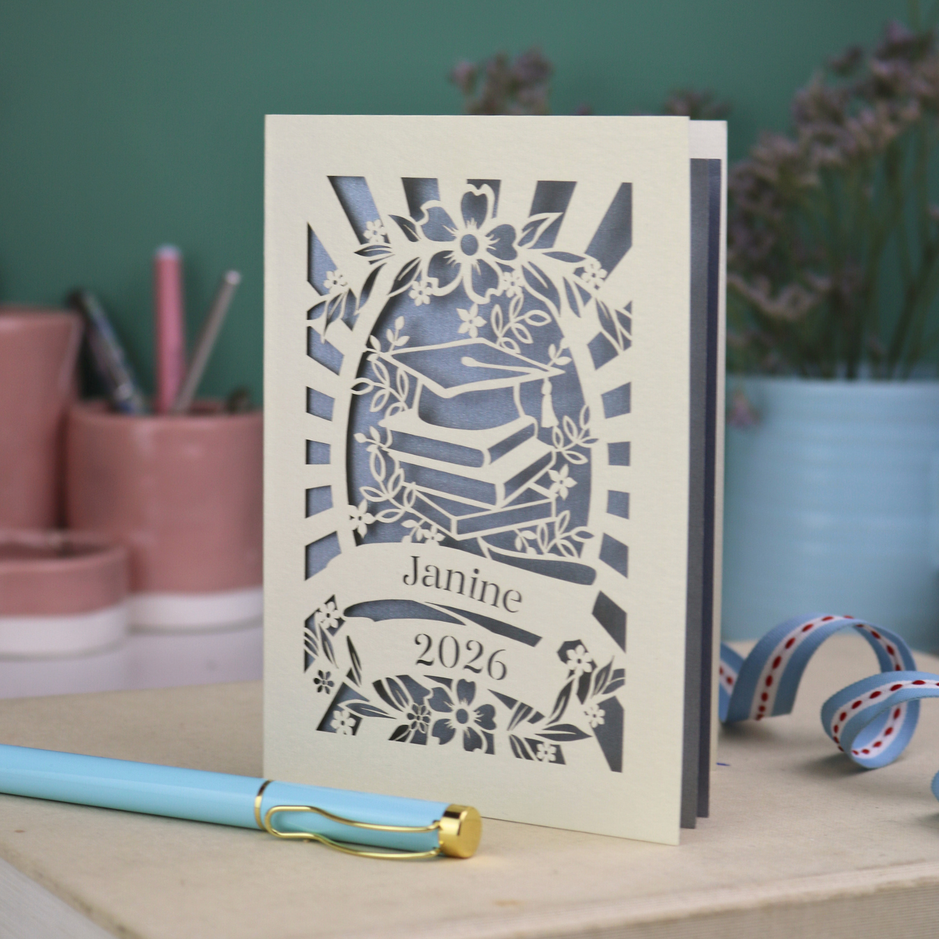 Personalised graduation card with cap and book design, and personalised with a name and the year in stencil font in a banner