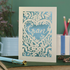 Decorative card with floral heart design and 'Gran' text on a green surface with stationery items.