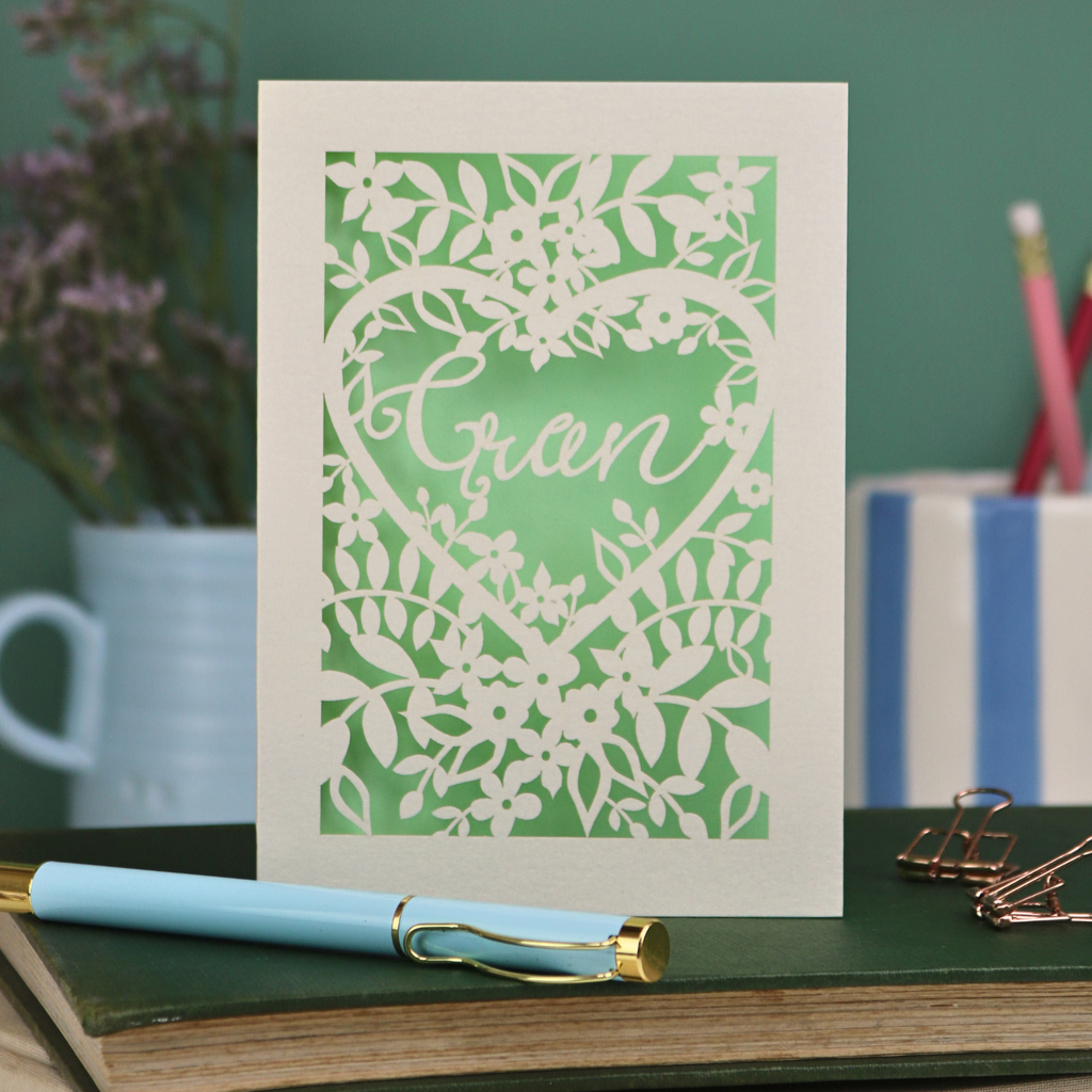 Laser cut Card with floral design and 'Gran' text on a green background, placed on a desk with stationery items.