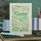 Laser cut Card with floral design and 'Gran' text on a green background, placed on a desk with stationery items.