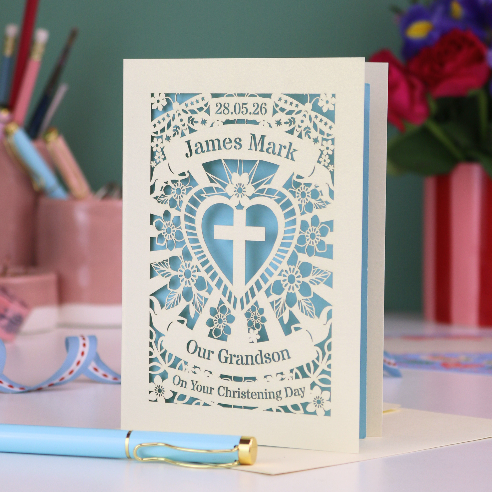 Personalized Grandson christening card with blue heart design and text, placed on a surface with stationery items in the background.