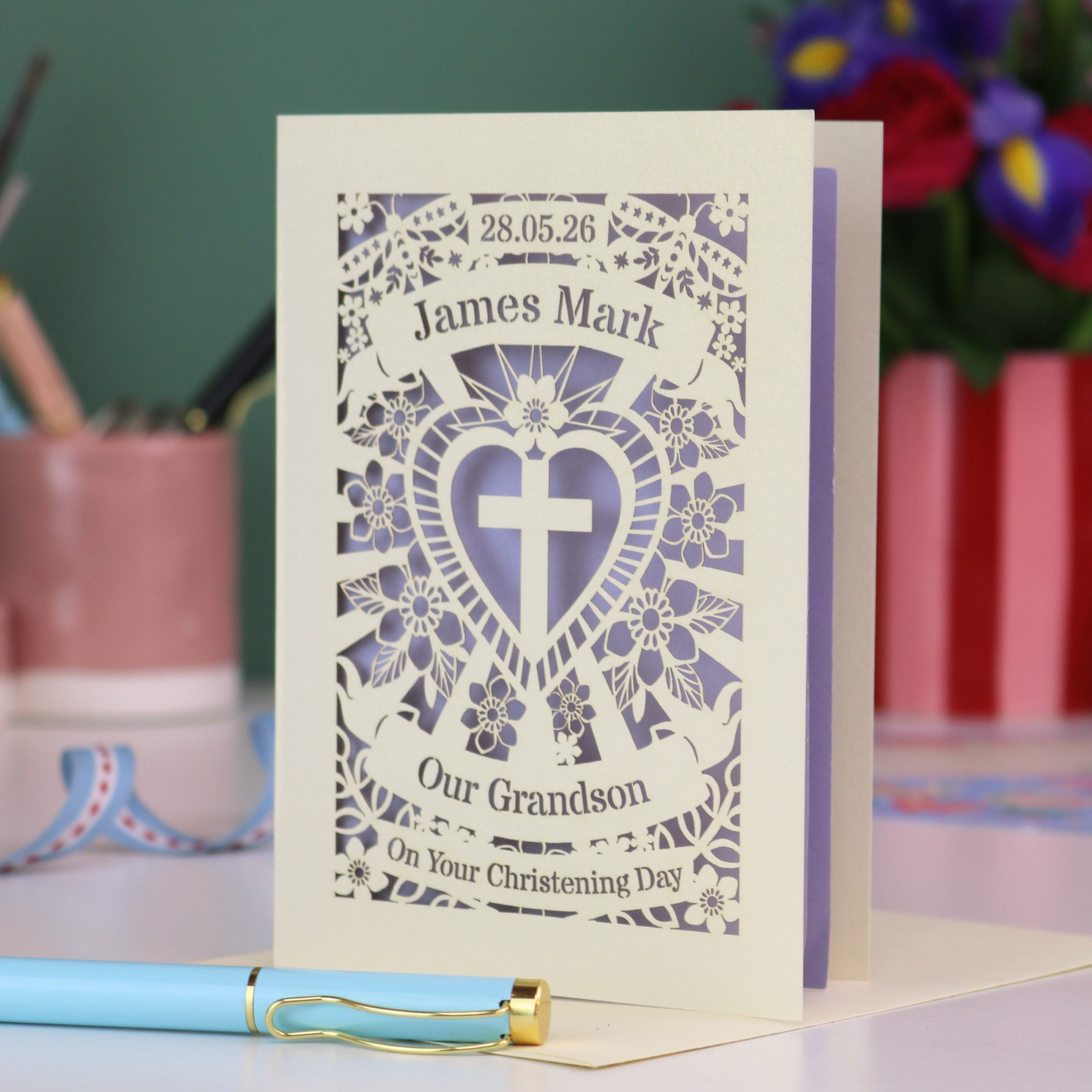 Personalized Grandson christening card with blue heart design and text, placed on a surface with stationery items in the background.