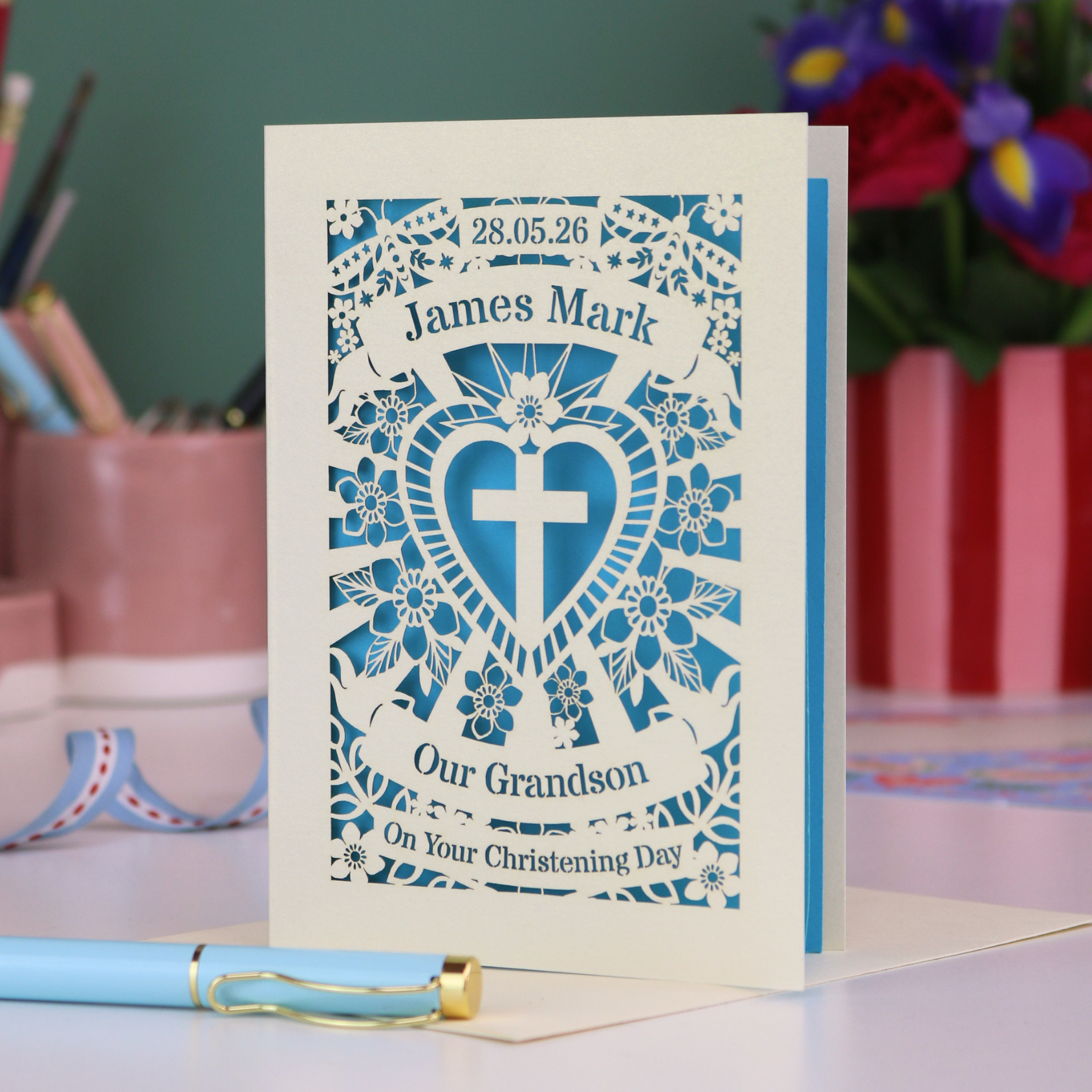 Personalized Grandson christening card with blue heart design and text, placed on a surface with stationery items in the background.