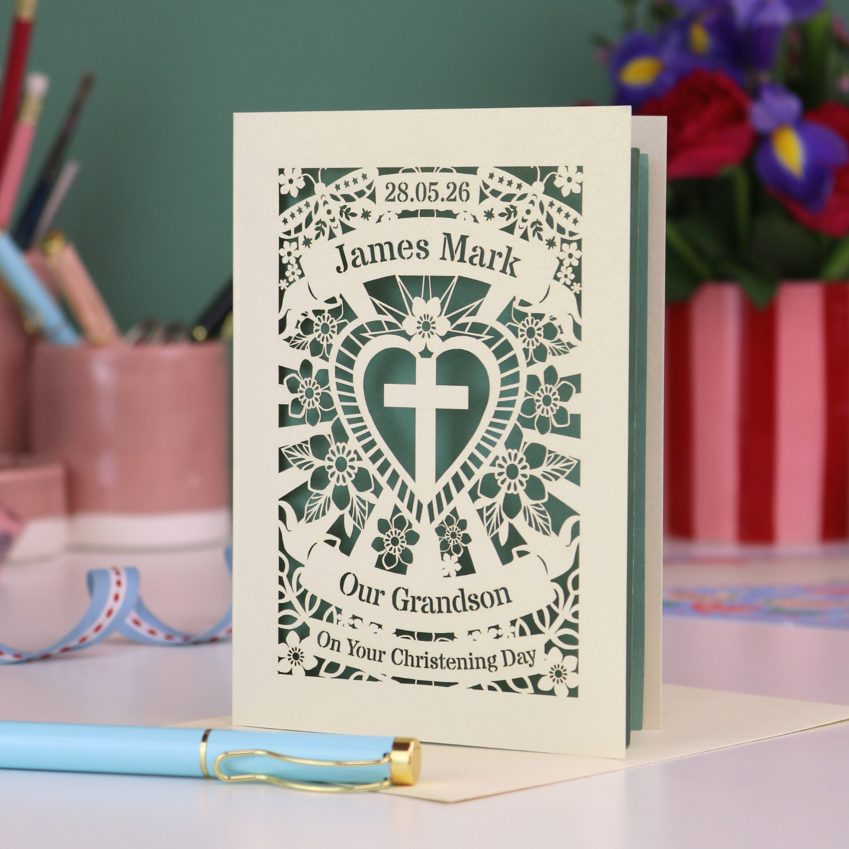 Personalized Grandson christening card with blue heart design and text, placed on a surface with stationery items in the background.