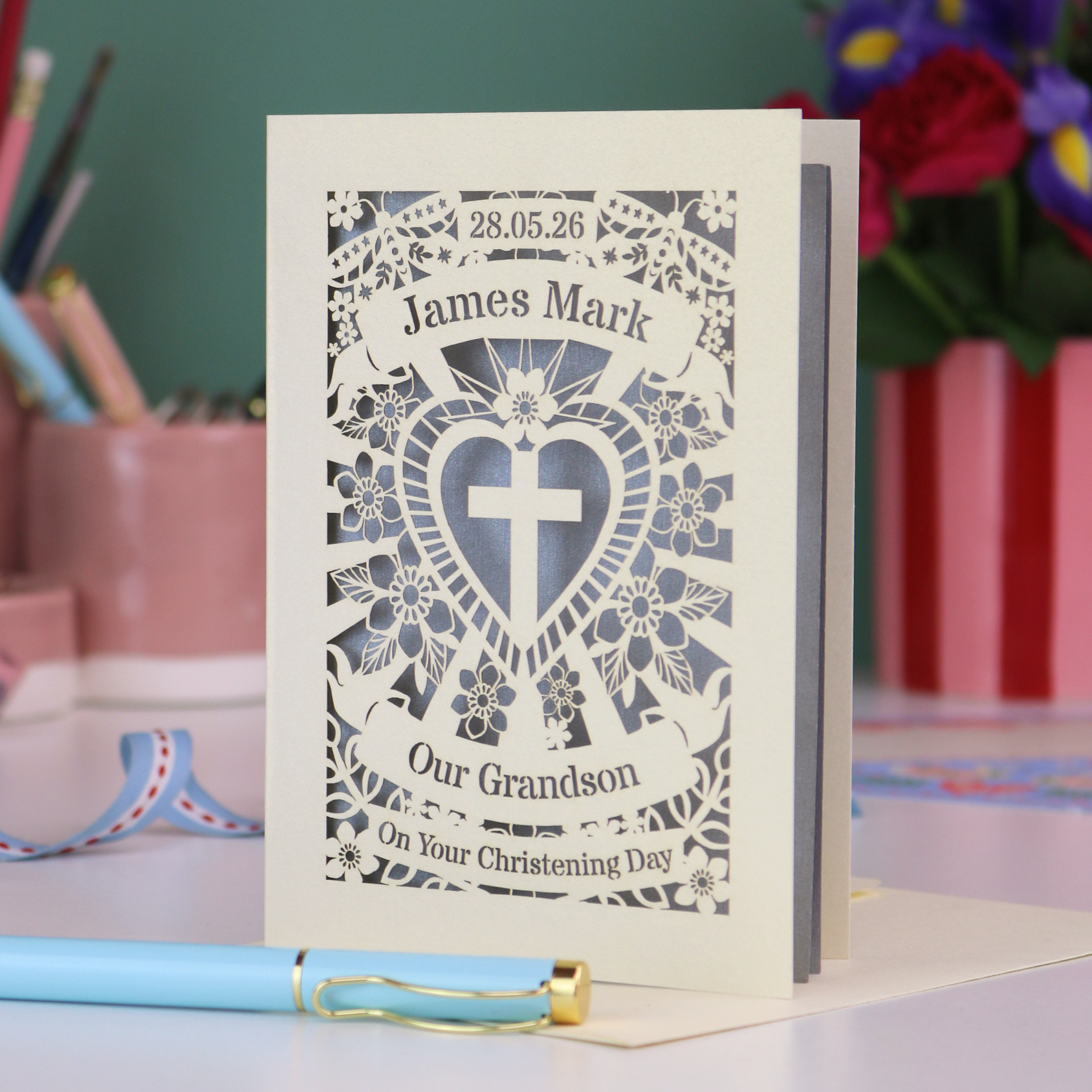 Personalized Grandson christening card with blue heart design and text, placed on a surface with stationery items in the background.