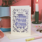 A laser cut Mother's Day card on white desk with stationery items and candles on pink background.