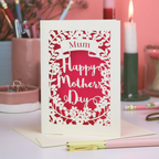 A laser cut Mothering Sunday card that says "mum, Happy Mother's 
Day". Card is on a white desk with stationery items and candles.