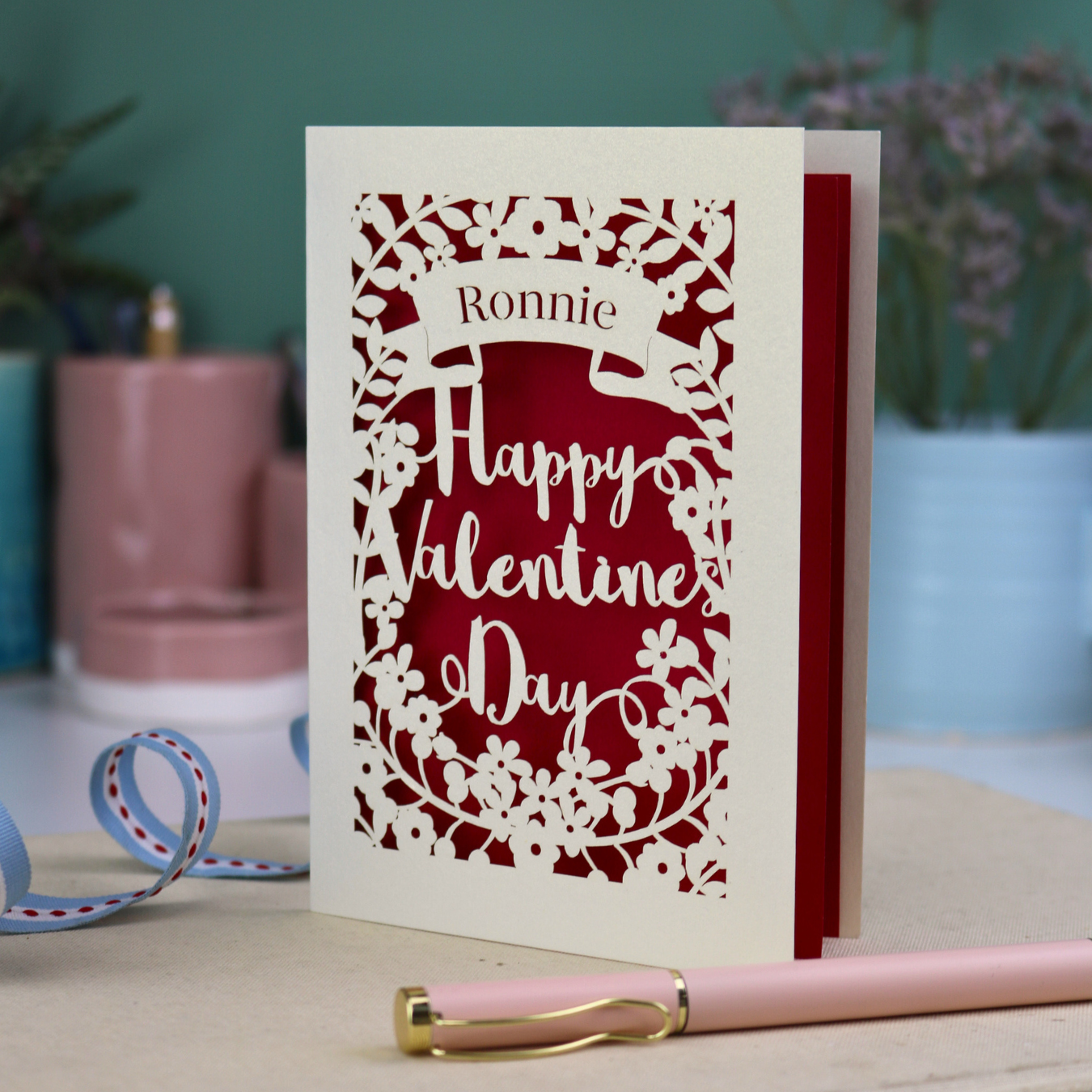 A personalised Valentine's Card with laser cut floral design, personalised with a name cut out in stencil font in a banner, with the text, "Happy Valentine's Day" in a script font.