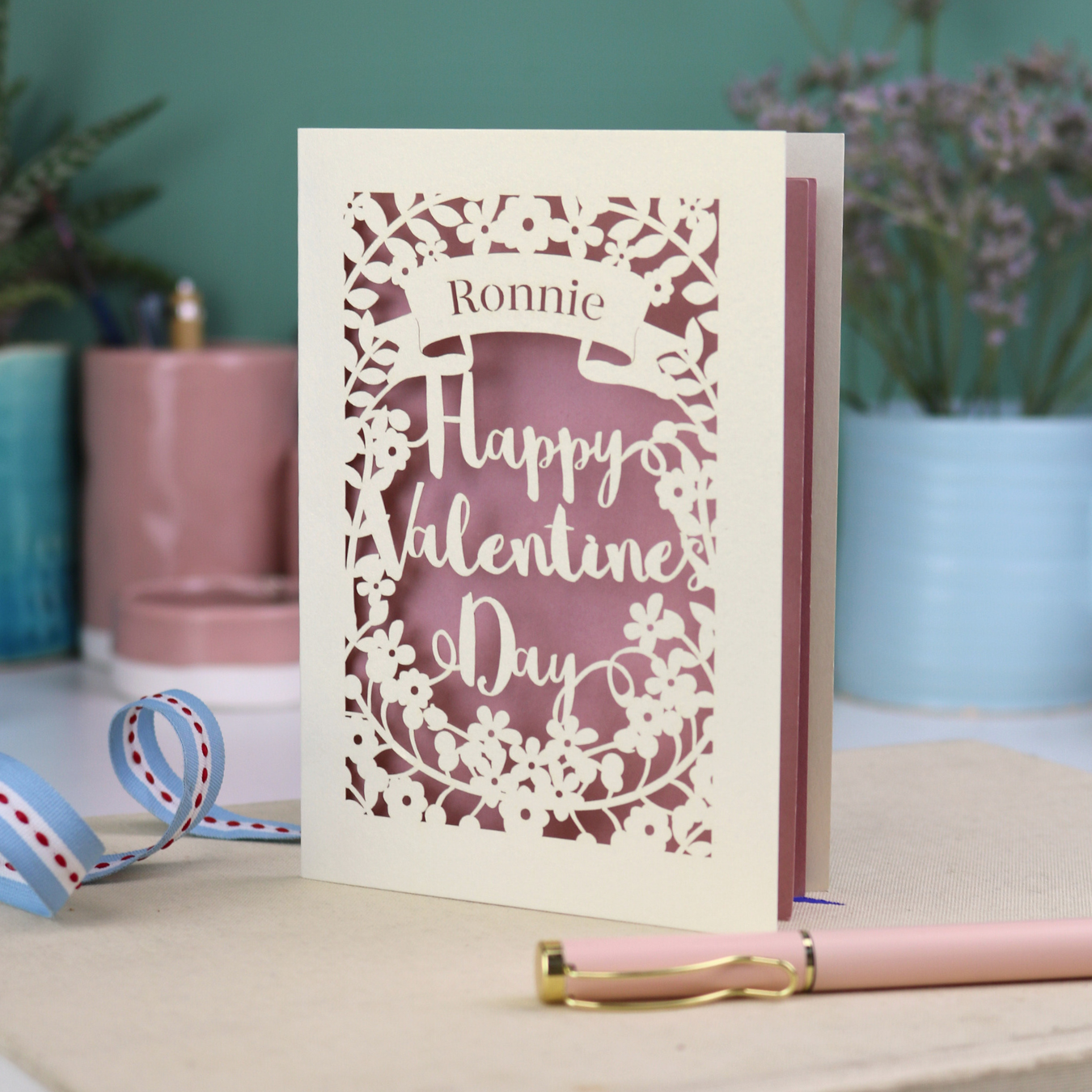 A personalised Valentine's Card with laser cut floral design, personalised with a name cut out in stencil font in a banner, with the text, "Happy Valentine's Day" in a script font.