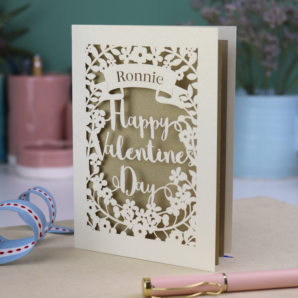 A personalised Valentine's Card with laser cut floral design, personalised with a name cut out in stencil font in a banner, with the text, "Happy Valentine's Day" in a script font.