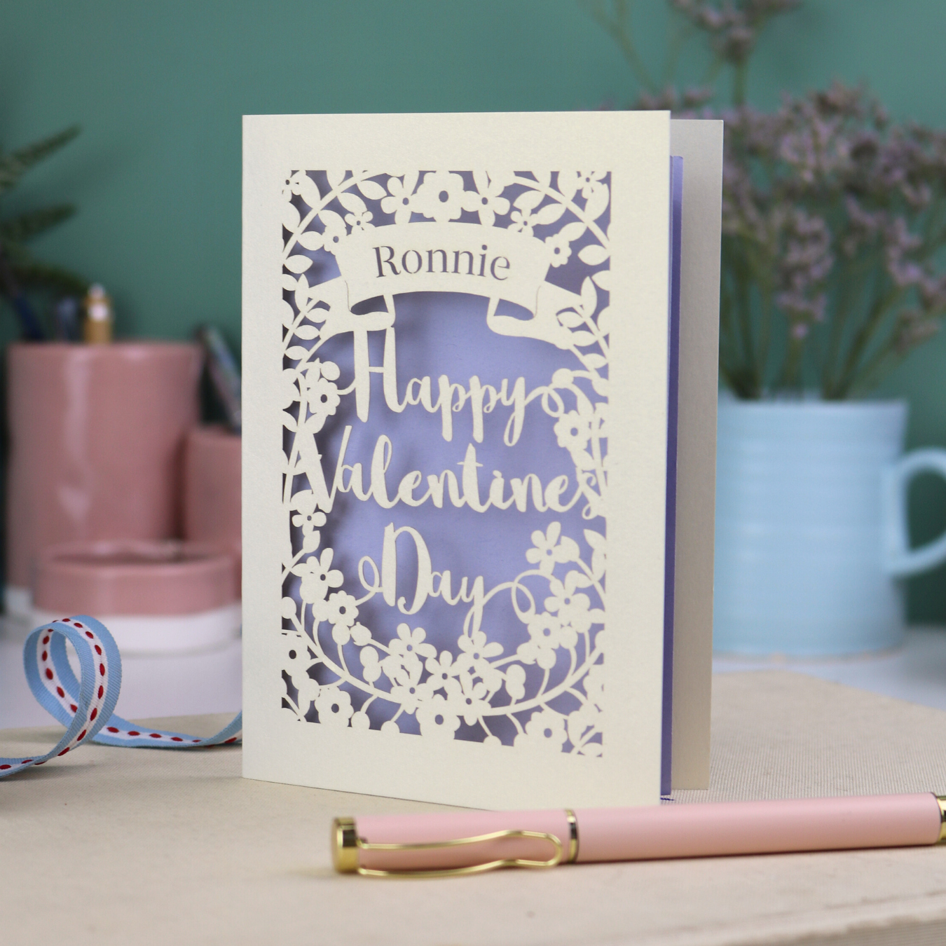 A personalised Valentine's Card with laser cut floral design, personalised with a name cut out in stencil font in a banner, with the text, "Happy Valentine's Day" in a script font.