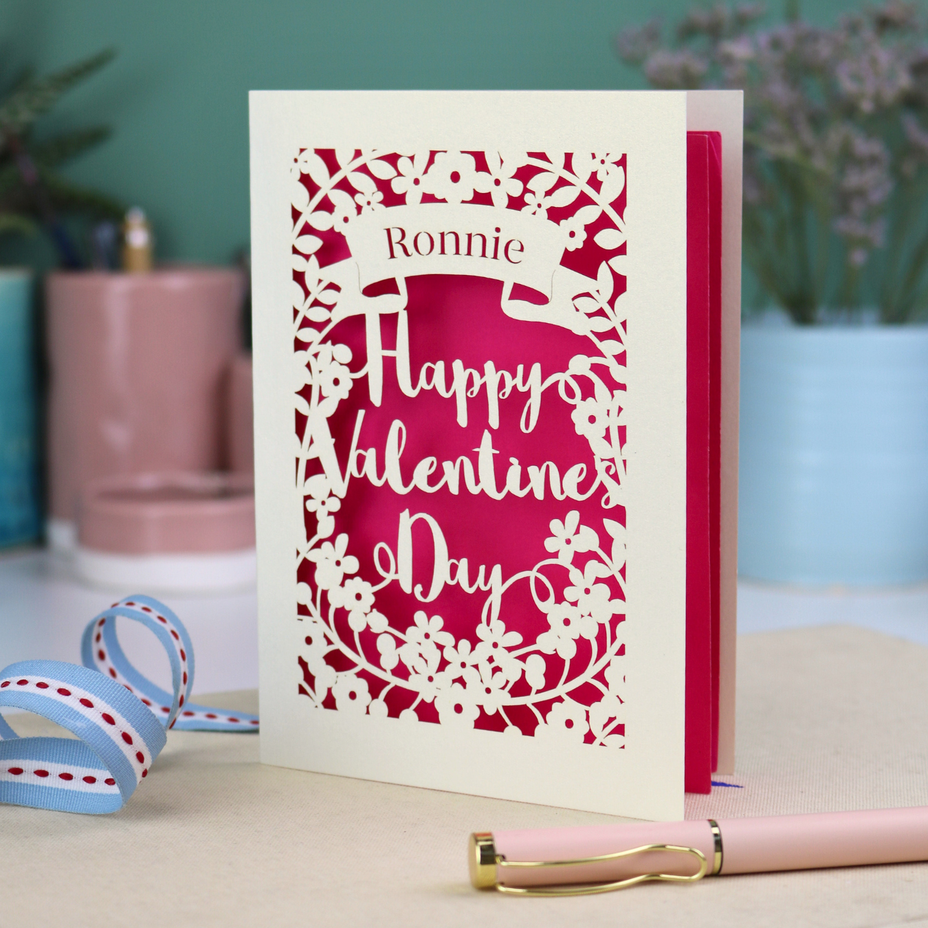 A personalised Valentine's Card with laser cut floral design, personalised with a name cut out in stencil font in a banner, with the text, "Happy Valentine's Day" in a script font.