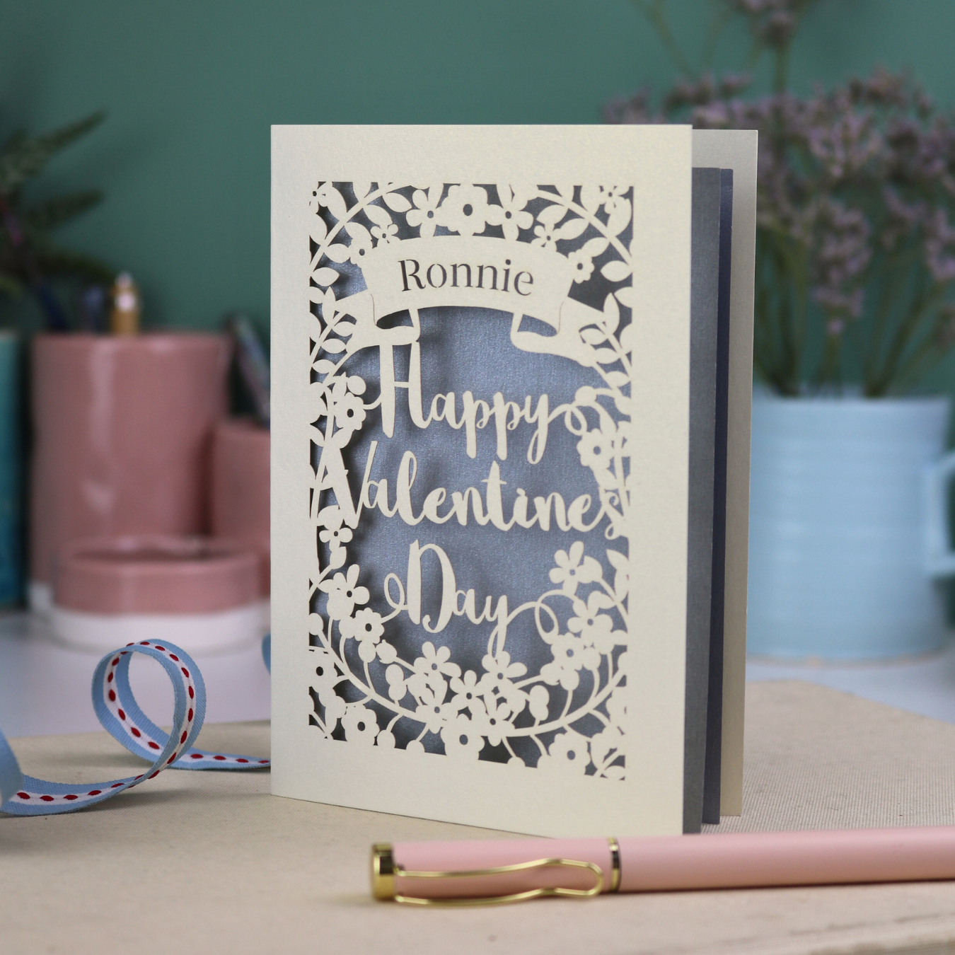 A personalised Valentine's Card with laser cut floral design, personalised with a name cut out in stencil font in a banner, with the text, "Happy Valentine's Day" in a script font.
