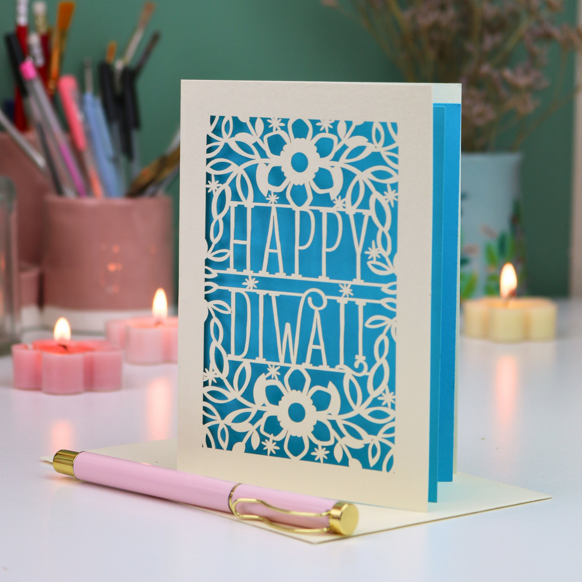 A handmade 'Happy Diwali' greeting card with a cream-colored laser-cut floral border and central text, displayed on a white desk with a pink pen in the foreground and blurred candles and stationery in the background.