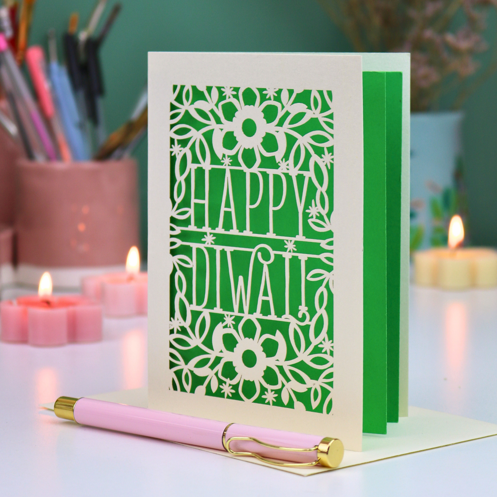A green and cream 'Happy Diwali' greeting card with a floral cut out design, displayed on a white surface with a pink and gold pen in front. The background features soft-focus pink candles, a cup of coloured pencils, and a teal wall.