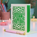 A green and cream 'Happy Diwali' greeting card with a floral cut out design, displayed on a white surface with a pink and gold pen in front. The background features soft-focus pink candles, a cup of coloured pencils, and a teal wall.
