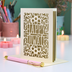 A laser-cut Diwali greeting card with 'HAPPY DIWALI' in the center and floral cutout patterns, displayed on a white surface with a pink pen in front and lit candles in the background.