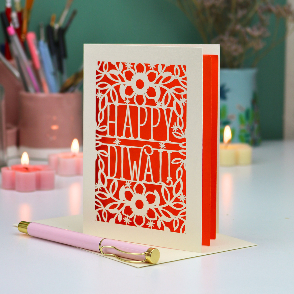 A cream-colored Diwali greeting card with a laser-cut lotus and floral design, standing upright on a desk with a pink pen resting in front. The card features cut out text 'Happy Diwali'