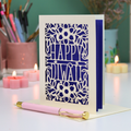 A handmade greeting card with a laser-cut Diwali design, standing on a white surface with a pink pen in front. The card features a floral pattern with the words 'HAPPY Diwali'.