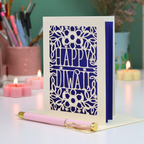 A handmade greeting card with a laser-cut Diwali design, standing on a white surface with a pink pen in front. The card features a floral pattern with the words 'HAPPY Diwali'.