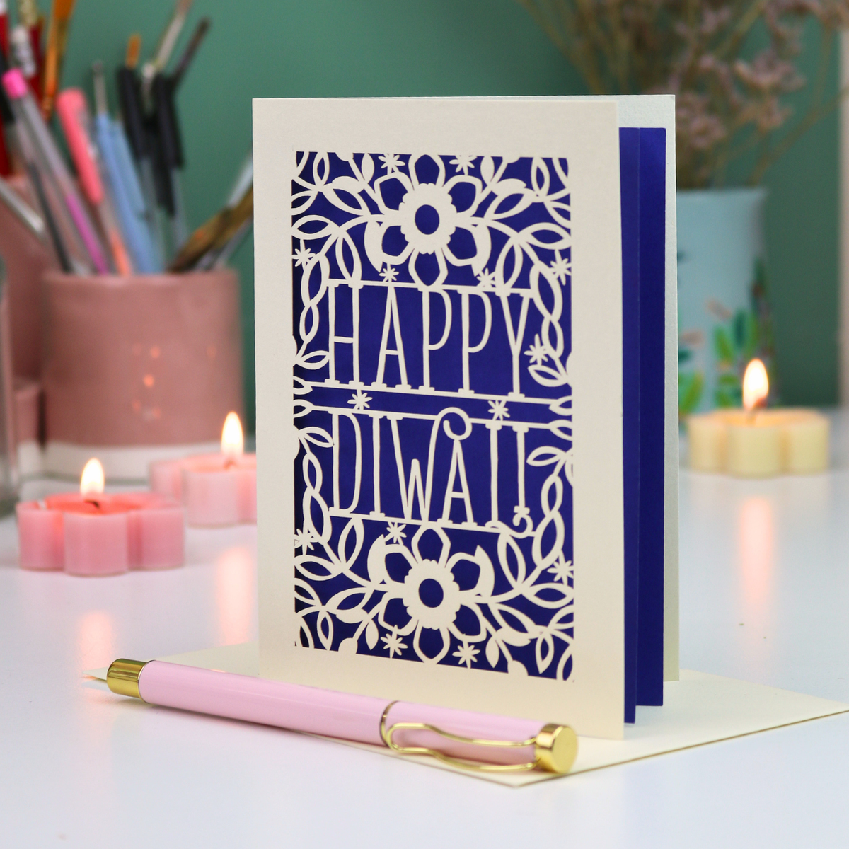 A handmade greeting card with a laser-cut Diwali design, standing on a white surface with a pink pen in front. The card features a floral pattern with the words 'HAPPY Diwali'.