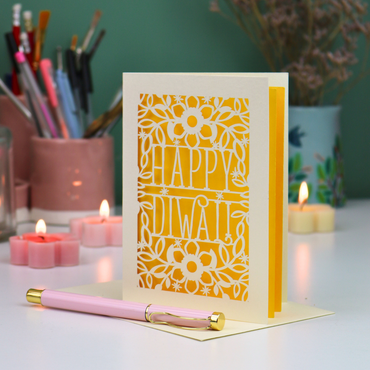 A decorative greeting card with a laser-cut floral border and the words “HAPPY DIWALI” in the centre, standing upright on a light surface. A pink pen with gold accents rests in front of the card.