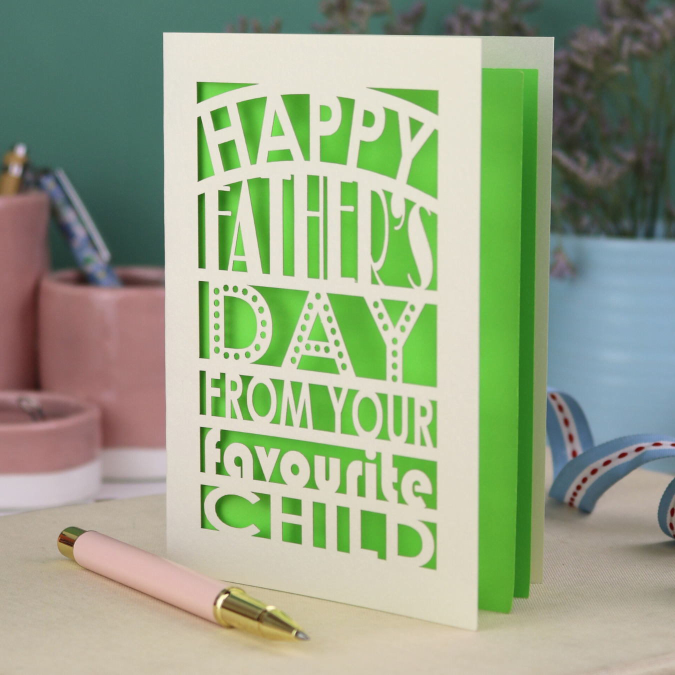 A funny Father's Day card with laser cut text, Happy Father's Day from your favourite child" on a table with a pen and decorative items.