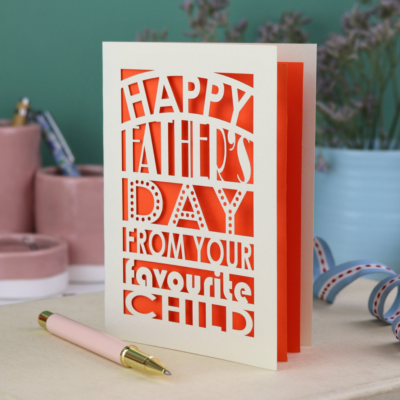 A funny Father's Day card with laser cut text, Happy Father's Day from your favourite child" on a table with a pen and decorative items.