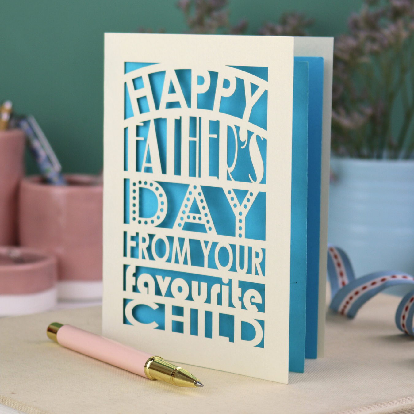 A funny Father's Day card with laser cut text, Happy Father's Day from your favourite child" on a table with a pen and decorative items.