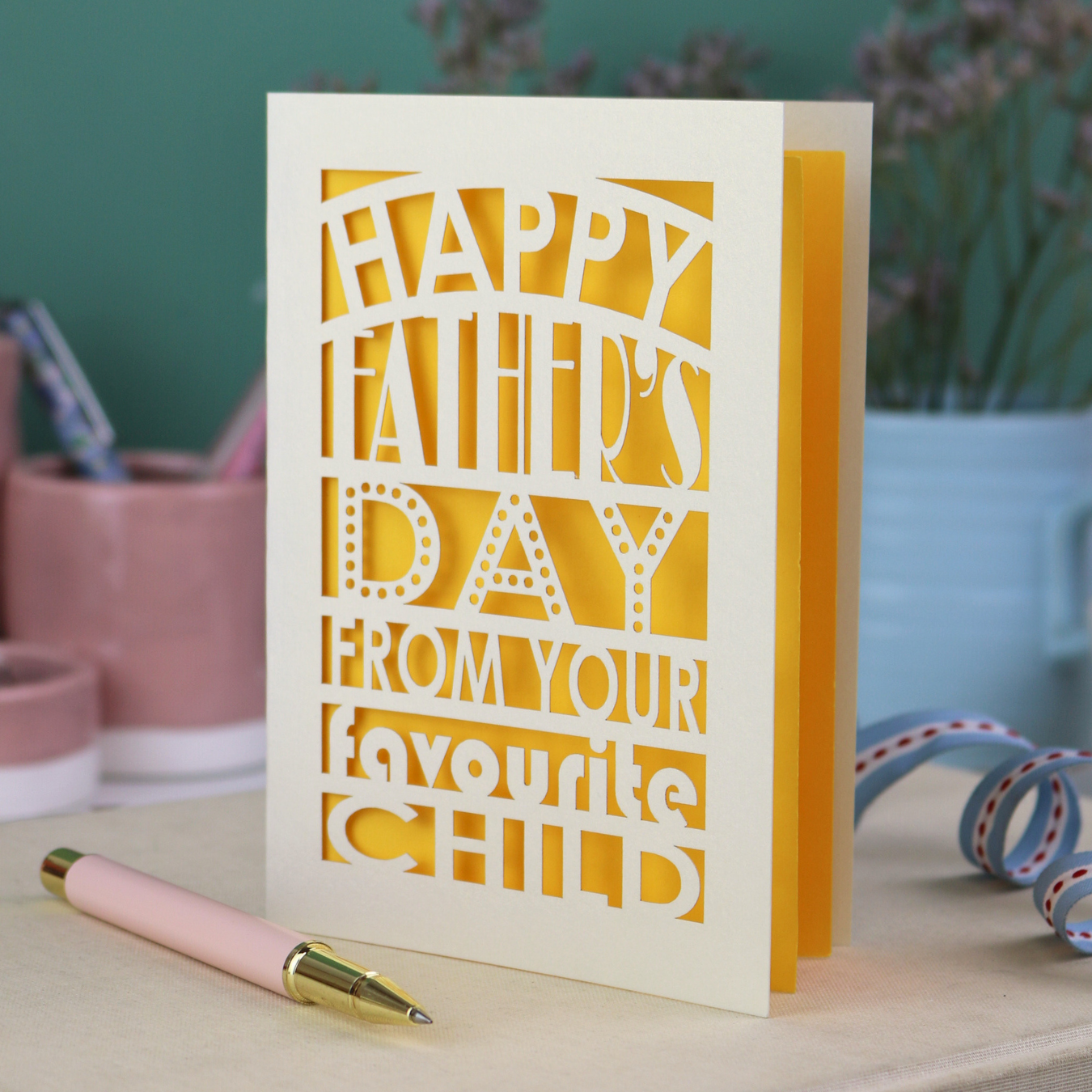 A funny Father's Day card with laser cut text, Happy Father's Day from your favourite child" on a table with a pen and decorative items.