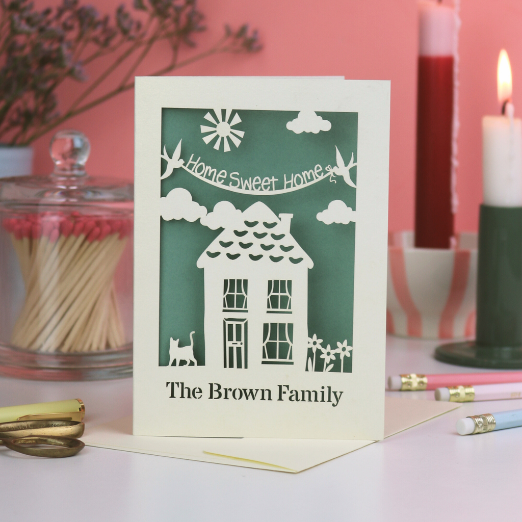 A laser cut housewarming card with house design and the words "home sweet home" with house design and personalised with the recipients' name. Card sists on white desk with pink background and stationery items