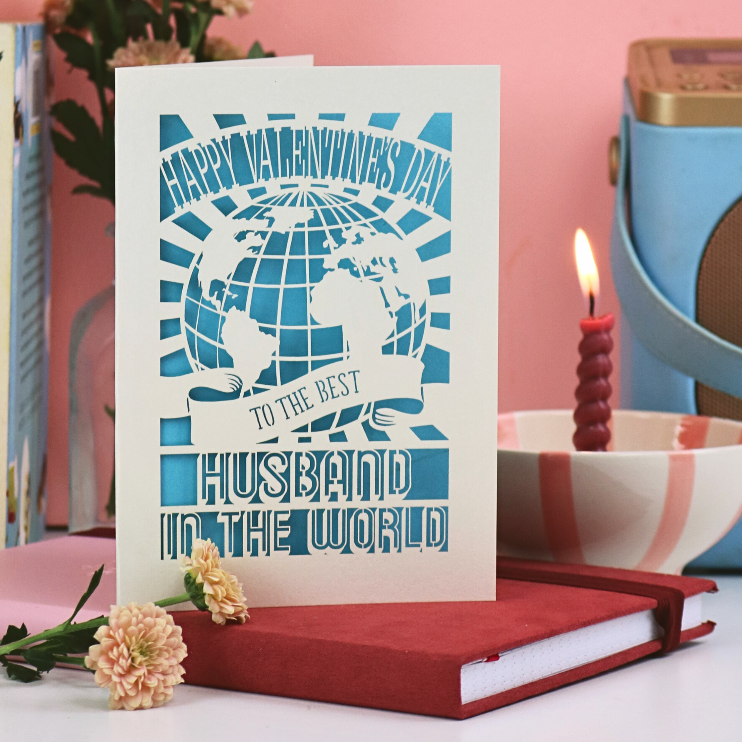 A laser cut husband Valentine card that says "Happy Valentine's Day to the best husband in the world", with globe design and blue backing, on a desk with flowers and candle. 