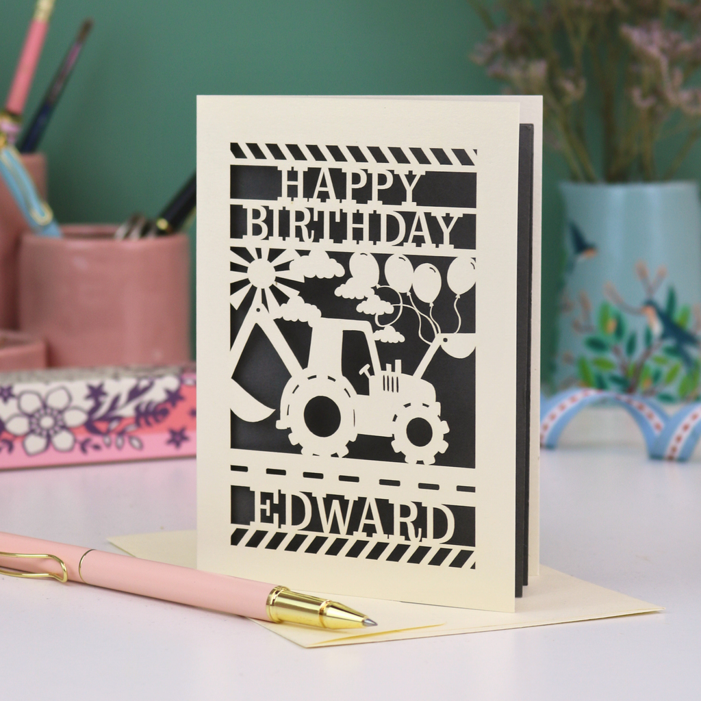 Personalised husband Birthday card with grey and cream design featuring a tractor and balloons, with 'Happy Birthday Edward' text, on a desk with stationery items.